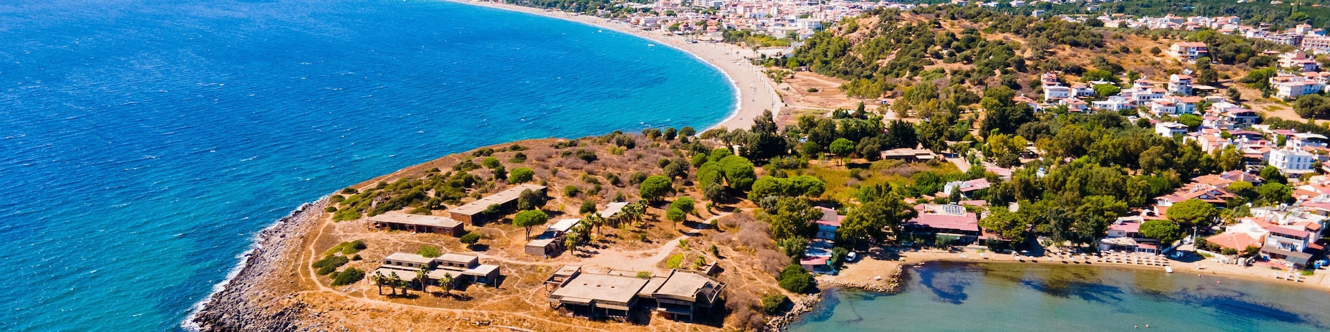 Vista aérea de un pueblo costero con una playa destacada, una pequeña isla con edificios y un telón de fondo montañoso.