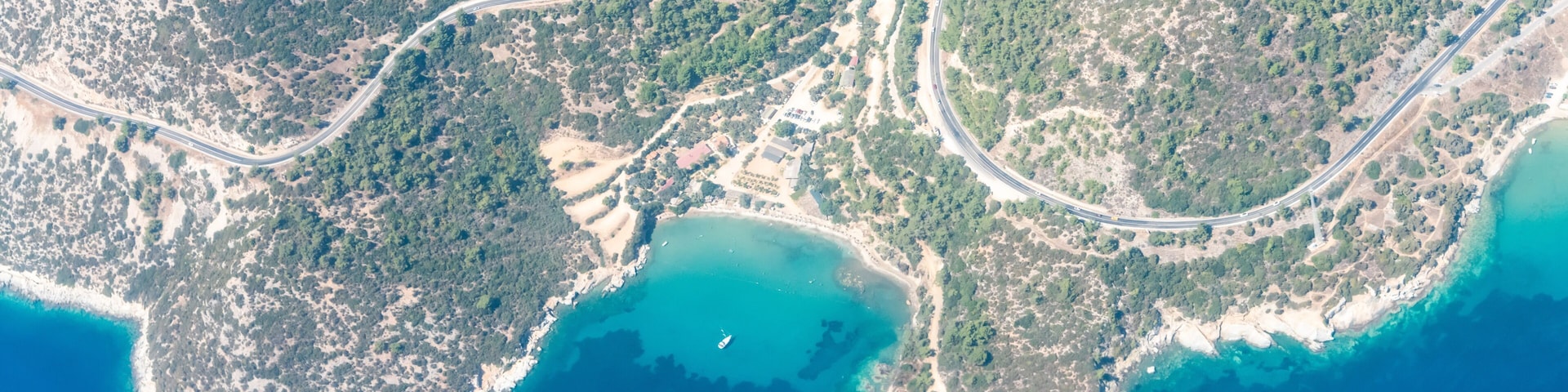 An aerial view of a coastal area with winding roads, dense forests, and a clear blue sea.
