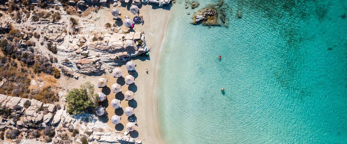 A rocky beach with clear turquoise water, umbrellas, and sparse vegetation.