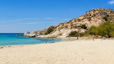 A sandy beach with clear turquoise water, rocky cliffs, and sparse vegetation.