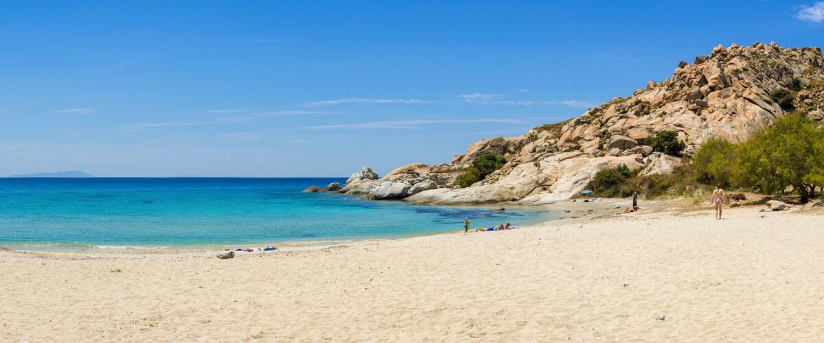 A sandy beach with clear turquoise water, rocky cliffs, and sparse vegetation.