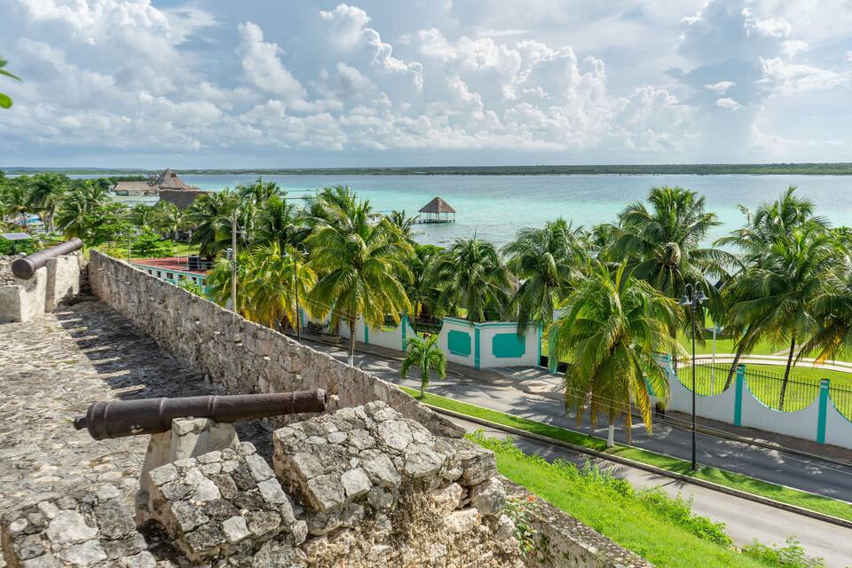 A coastal view with a stone fort, palm trees, a thatched-roof hut, and turquoise waters.