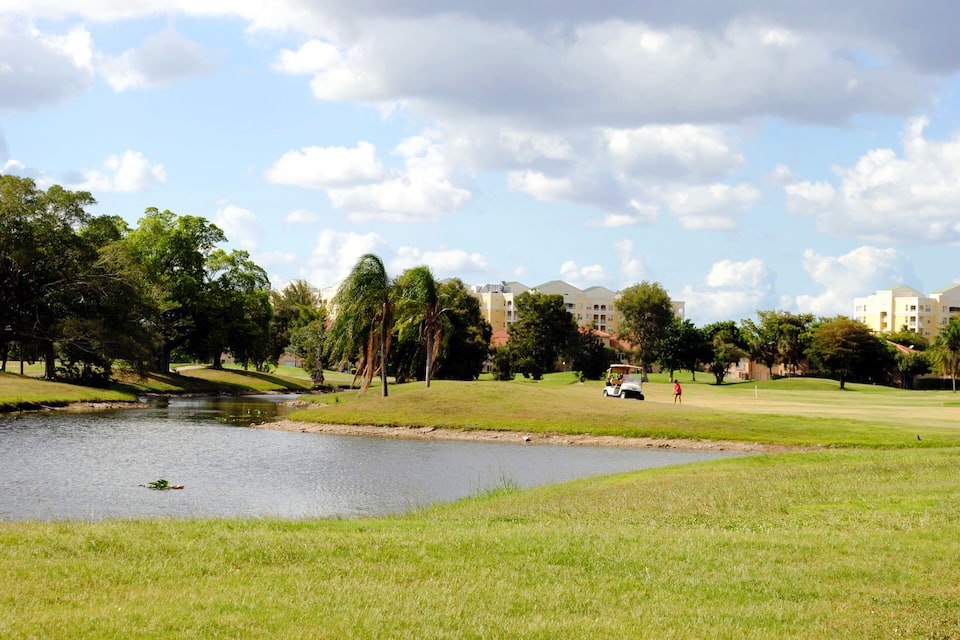 Un campo de golf con un estanque, césped verde, árboles y edificios al fondo.
