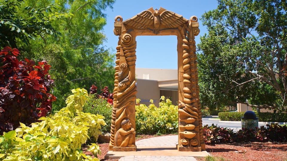 A stone archway with intricate carvings, surrounded by lush greenery and a building in the background.