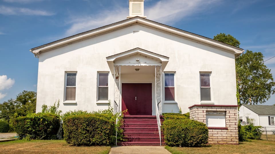 A white church with a red door and a brick foundation labeled "First Baptist Church".