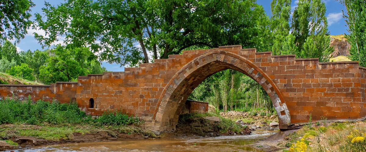 A brick bridge arching over a flowing river, surrounded by lush greenery and trees.