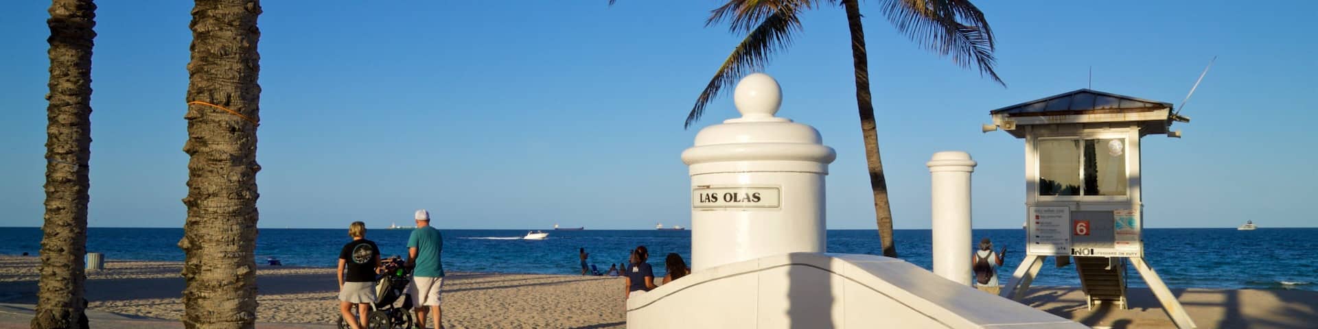 A beach with palm trees, a lifeguard tower, and people walking.
