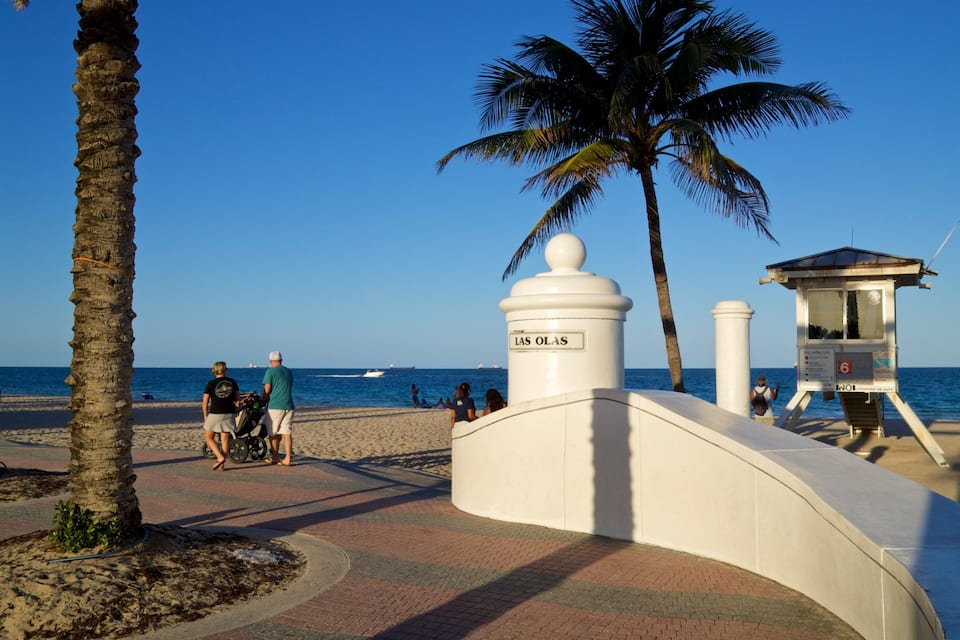Una playa con palmeras, una torre de salvavidas y gente caminando.