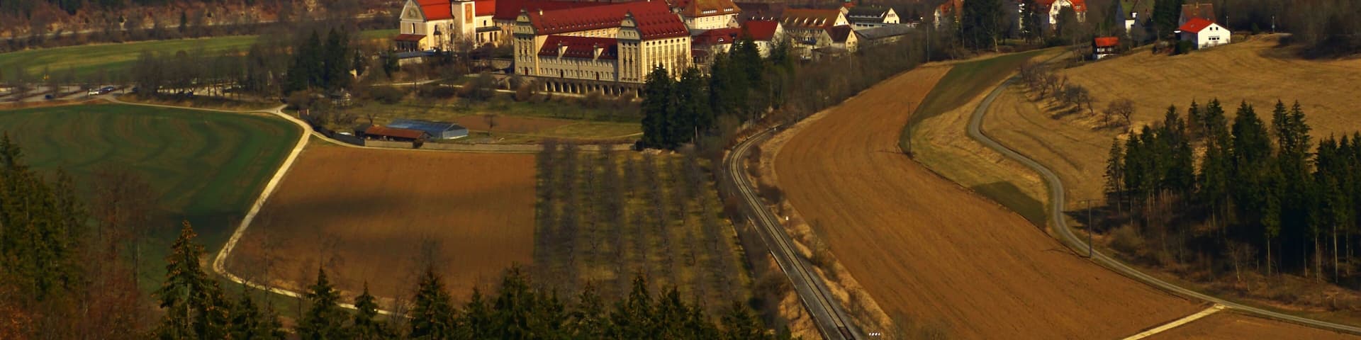 A rural landscape with a large estate featuring a red-roofed building, a church, and a railway track.