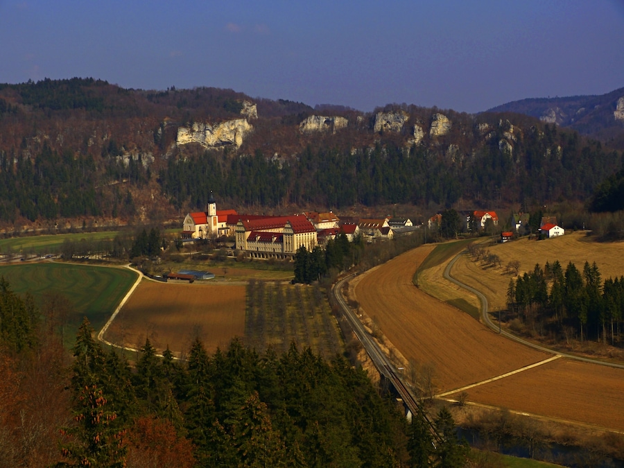 Paisaje rural con una gran propiedad que incluye un edificio de techo rojo, una iglesia y una vía de tren.