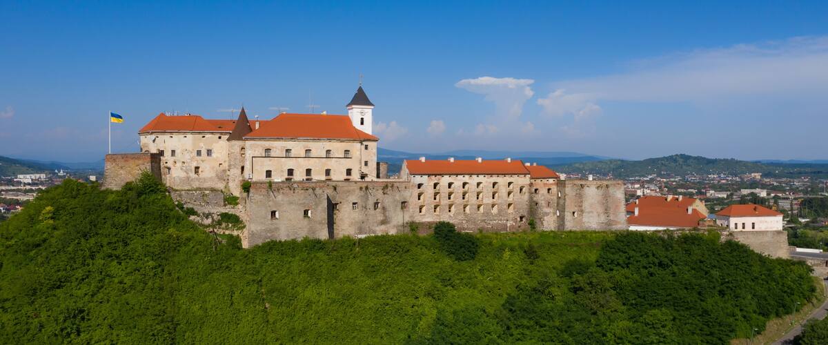 A historic castle on a hill with a red roof, surrounded by greenery and a clear sky.