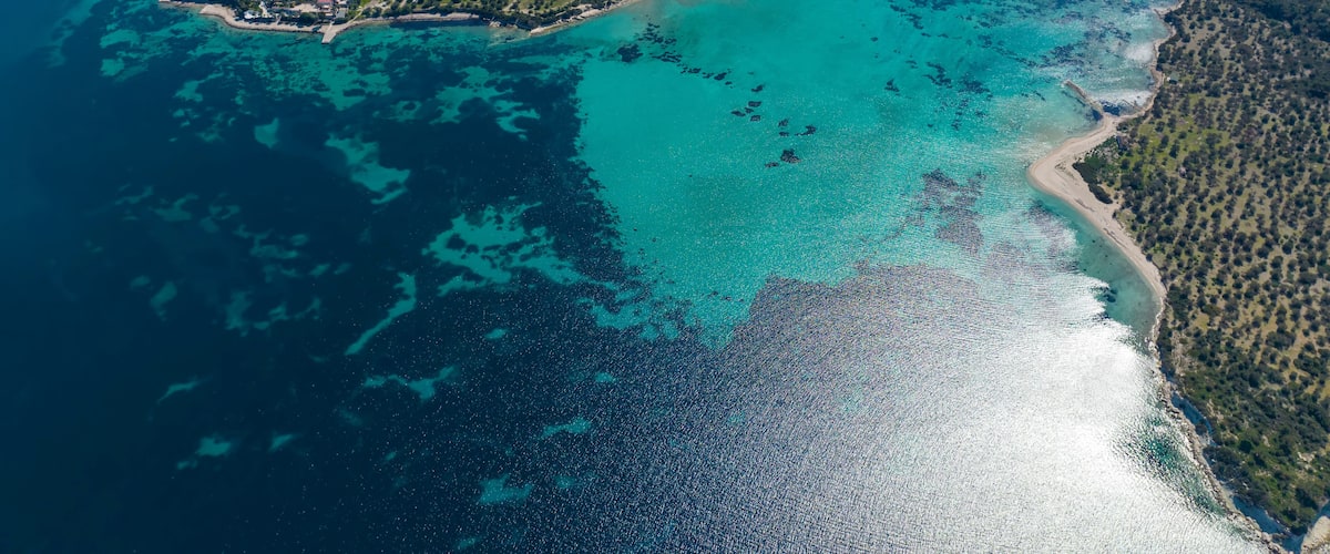 An aerial view of a coastal area with clear turquoise waters, a small island with buildings, and a forested peninsula.