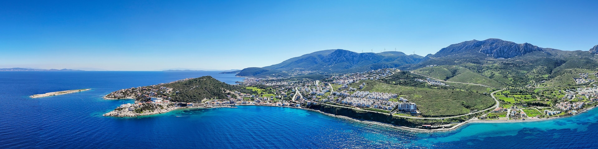 Un pueblo costero con montañas al fondo, aguas azules cristalinas y aerogeneradores.