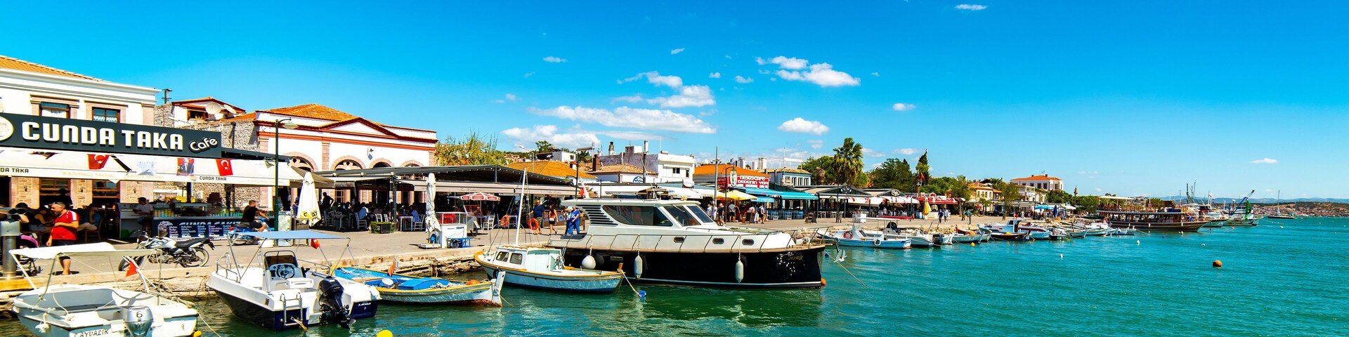 Un pueblo costero con un puerto deportivo, barcos y un café llamado 'Sunda Taka Cafe'.