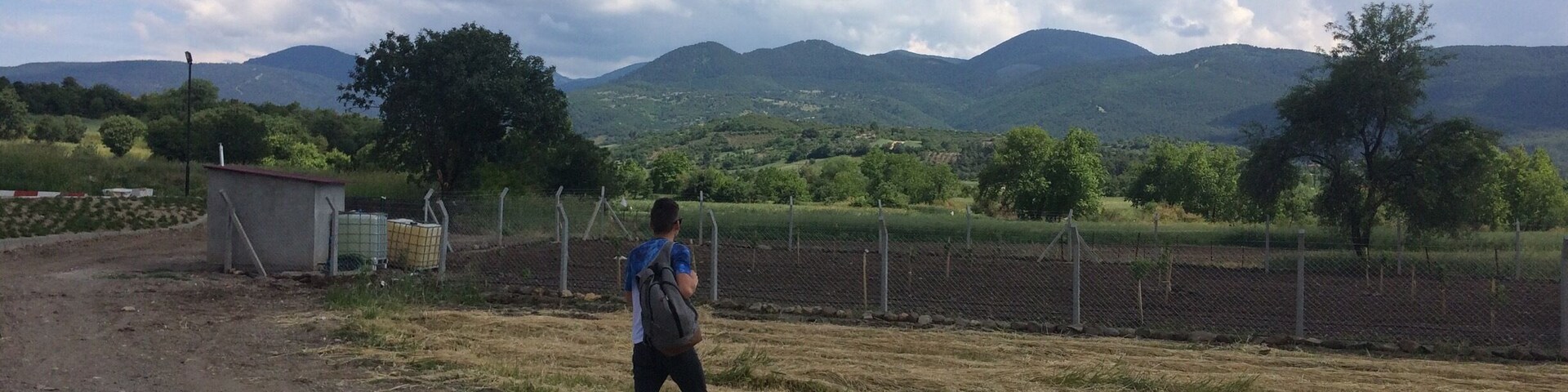 A rural landscape with a dirt path, a small building, a fenced field, and mountains in the background.