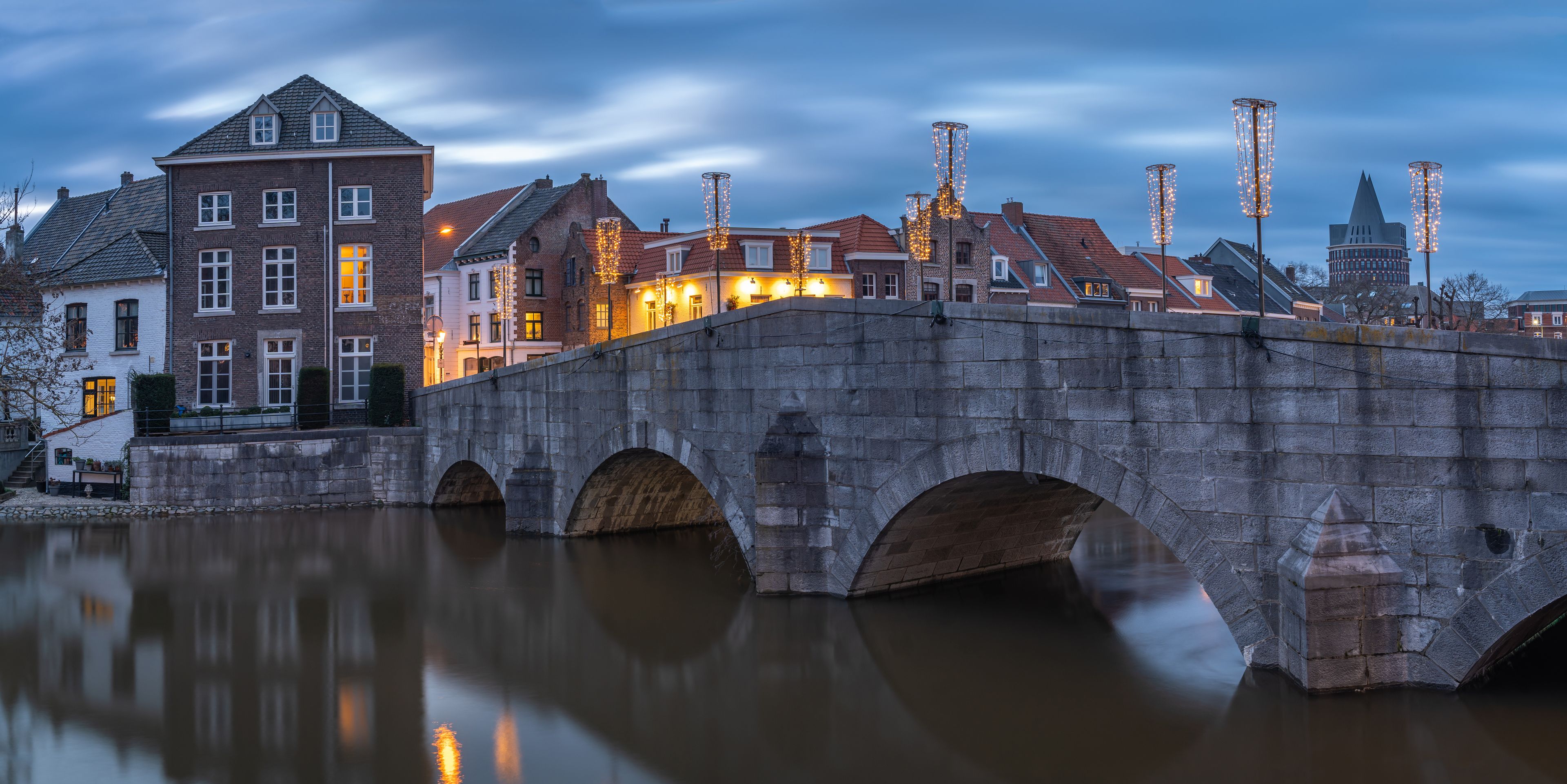 Un pont de pierre enjambant une rivière paisible, avec des bâtiments de chaque côté et des éclairages décoratifs.