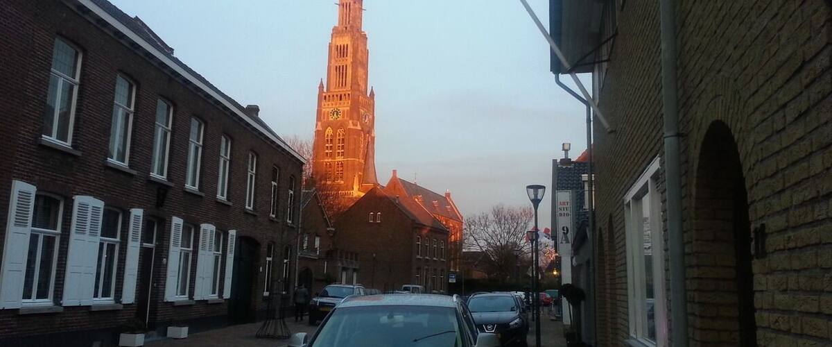 A street with brick buildings, parked cars, and a tall church tower in the background.