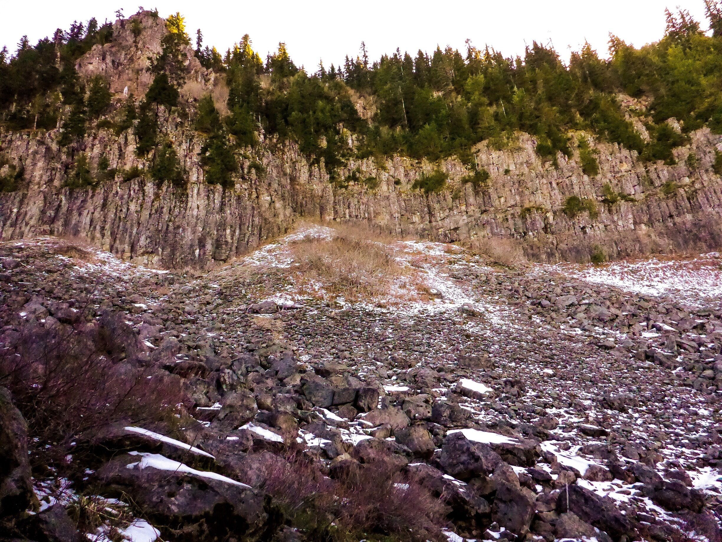 A rocky terrain with sparse snow patches and a forested cliff in the background.