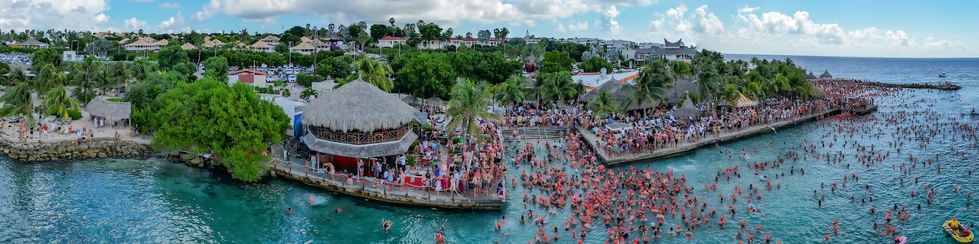 A crowded beach with people swimming and a thatched-roof structure.