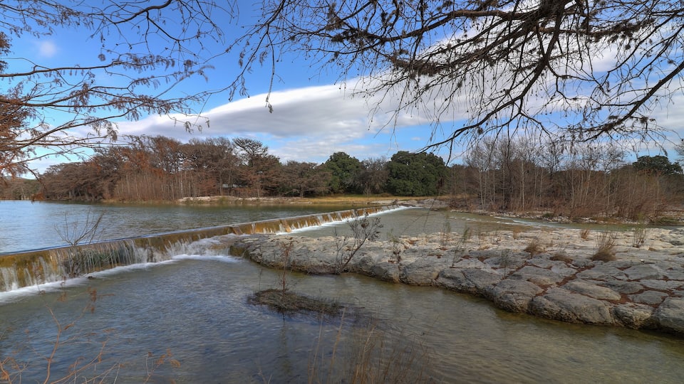 A river with a small waterfall, rocky banks, and trees in the background.
