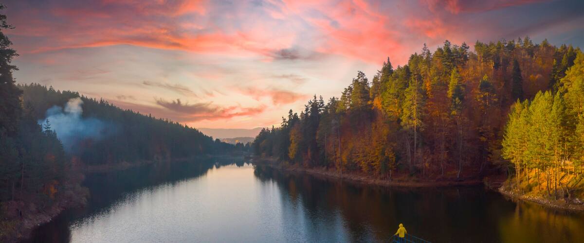 A calm river with a bridge and a person in yellow, surrounded by autumn-colored trees.