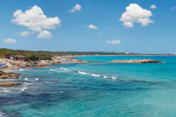 Paisagem costeira com águas turquesa cristalinas, costas rochosas, uma praia de areia e um farol ao longe.
