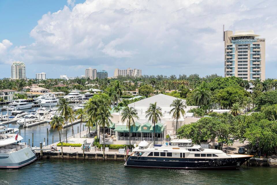A marina with yachts, a large building with a green roof, and palm trees.