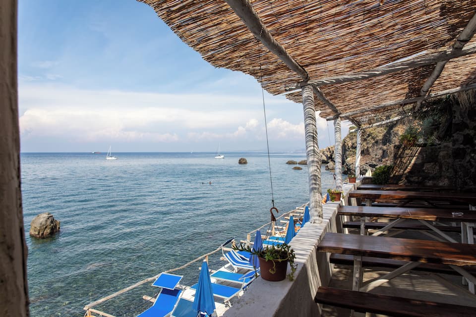 Une terrasse en bord de mer, avec des bancs en bois et des fauteuils bleus, d’où l’on peut admirer la mer et les bateaux qui naviguent au loin.