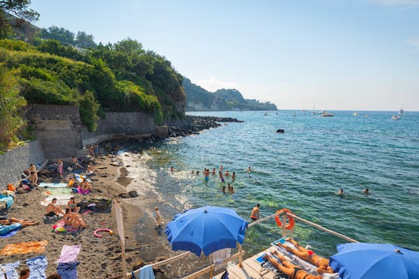A beach with people sunbathing and swimming, surrounded by greenery and rocky cliffs.
