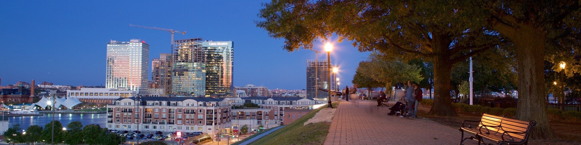 Paisagem urbana ao anoitecer com um rio, um parque com bancos e edifícios modernos.