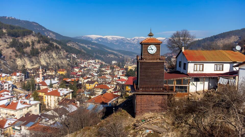 A clock tower with a red roof, surrounded by a town with snow-covered mountains in the background.