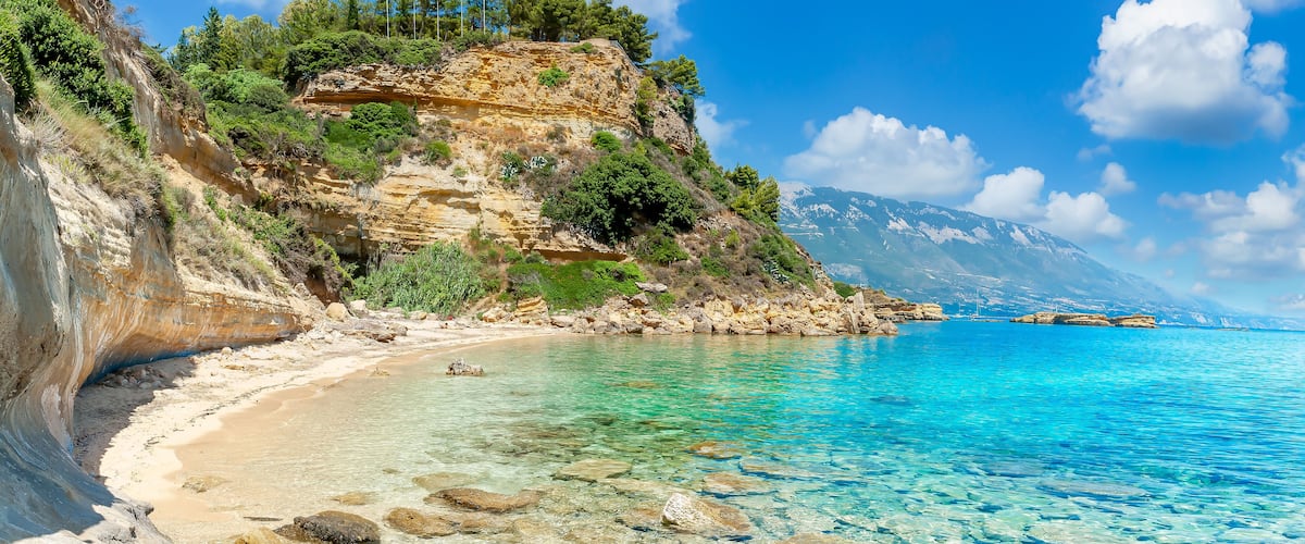A coastal landscape with clear turquoise waters, rocky cliffs, and a mountainous backdrop.
