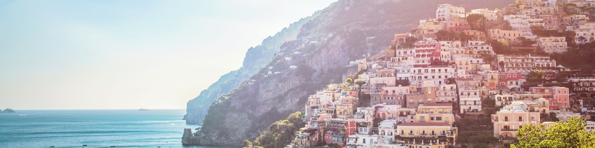 Un pueblo costero con edificios de colores en una colina, barcos en el agua y cielo despejado.