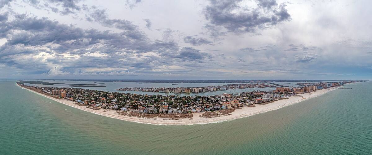 An aerial view of a coastal city with a beach, water bodies, and a cloudy sky.