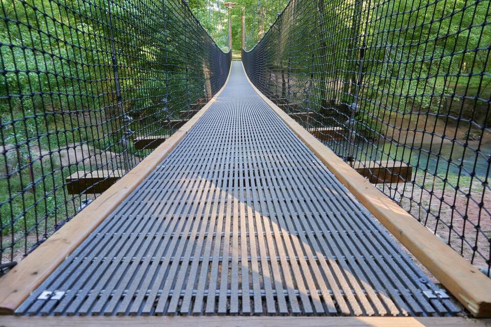 A suspension bridge with a metal grid floor, surrounded by dense greenery.