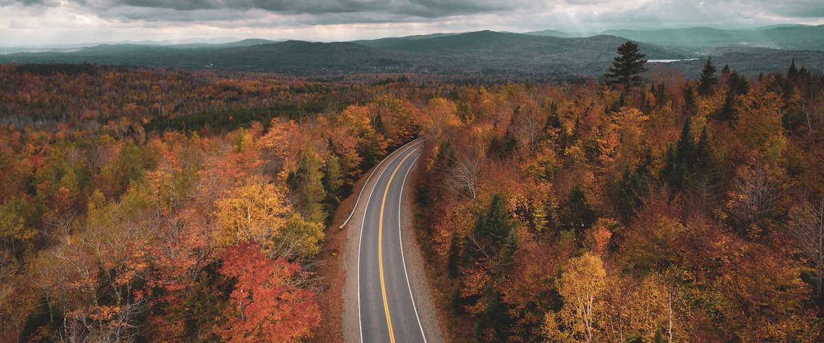 A winding road through a forest with autumn foliage, under a cloudy sky.