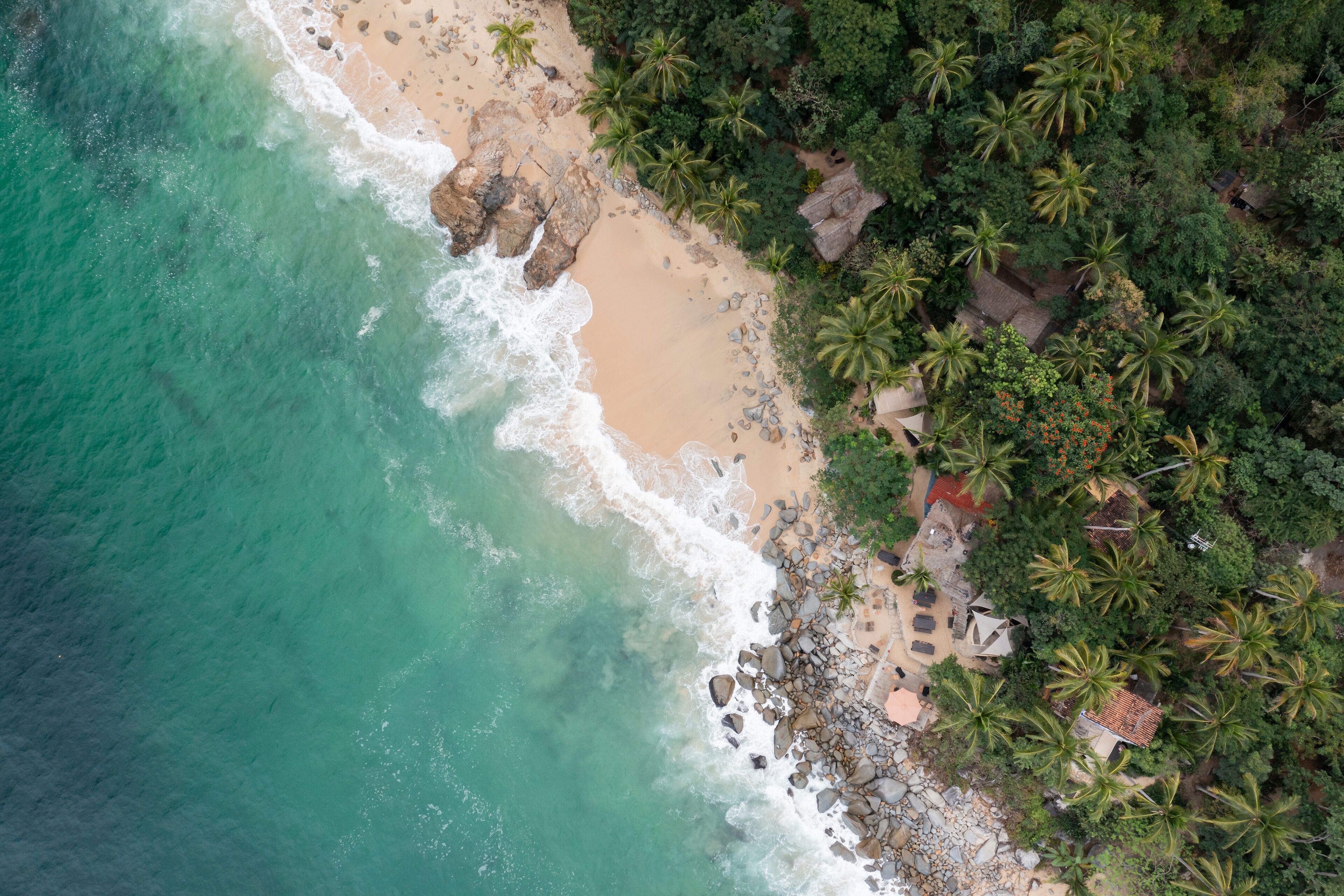 Aerial View of Casitas Marika beach, small beach huts and a thick cover of palm trees and forest. 