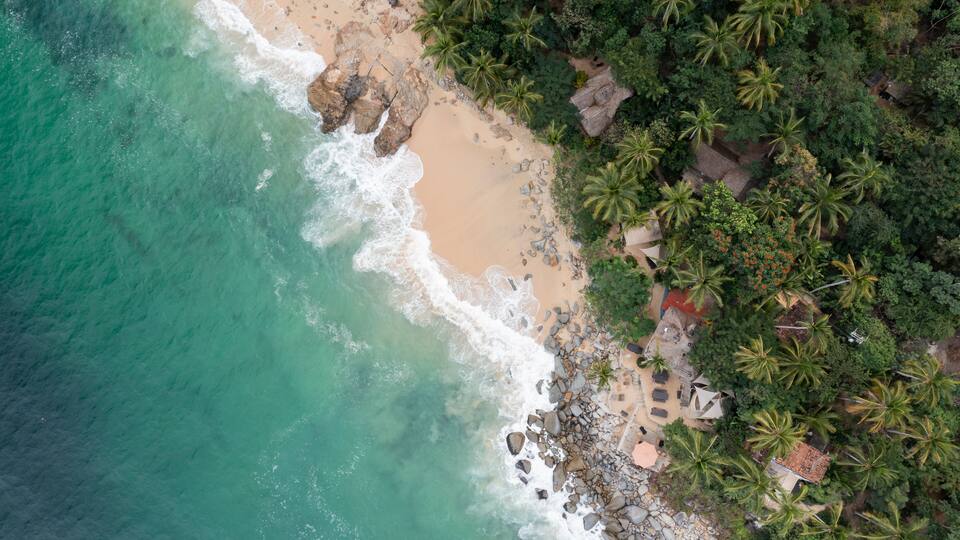 Aerial View of Casitas Marika beach, small beach huts and a thick cover of palm trees and forest.
