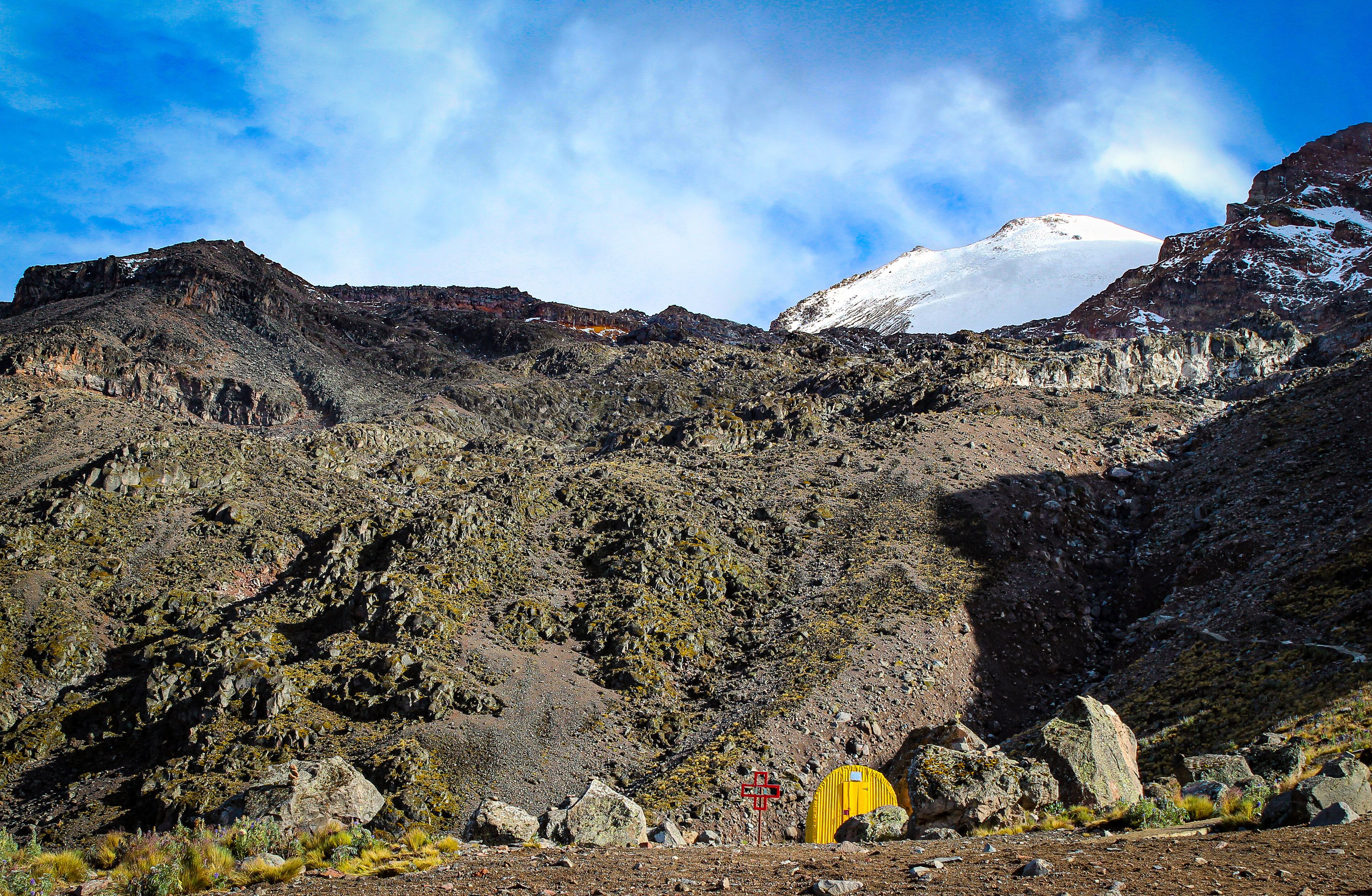 A rugged mountain landscape with rocky terrain, a snow-capped peak, and a yellow tent.