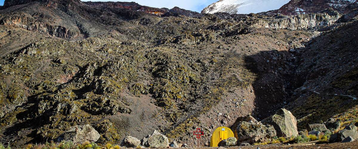 A rugged mountain landscape with rocky terrain, a snow-capped peak, and a yellow tent.