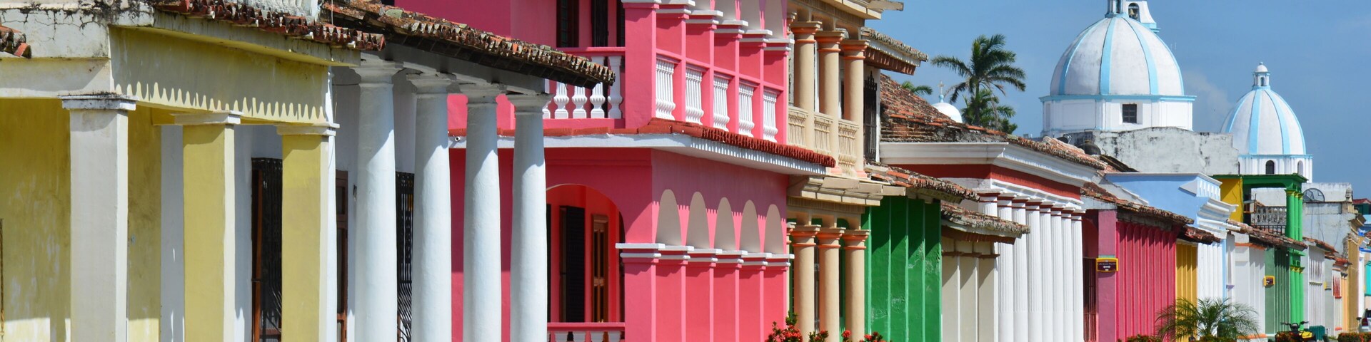 Una calle colorida bordeada de edificios de colores pastel, un cielo azul despejado y una iglesia con cúpulas azules al fondo.