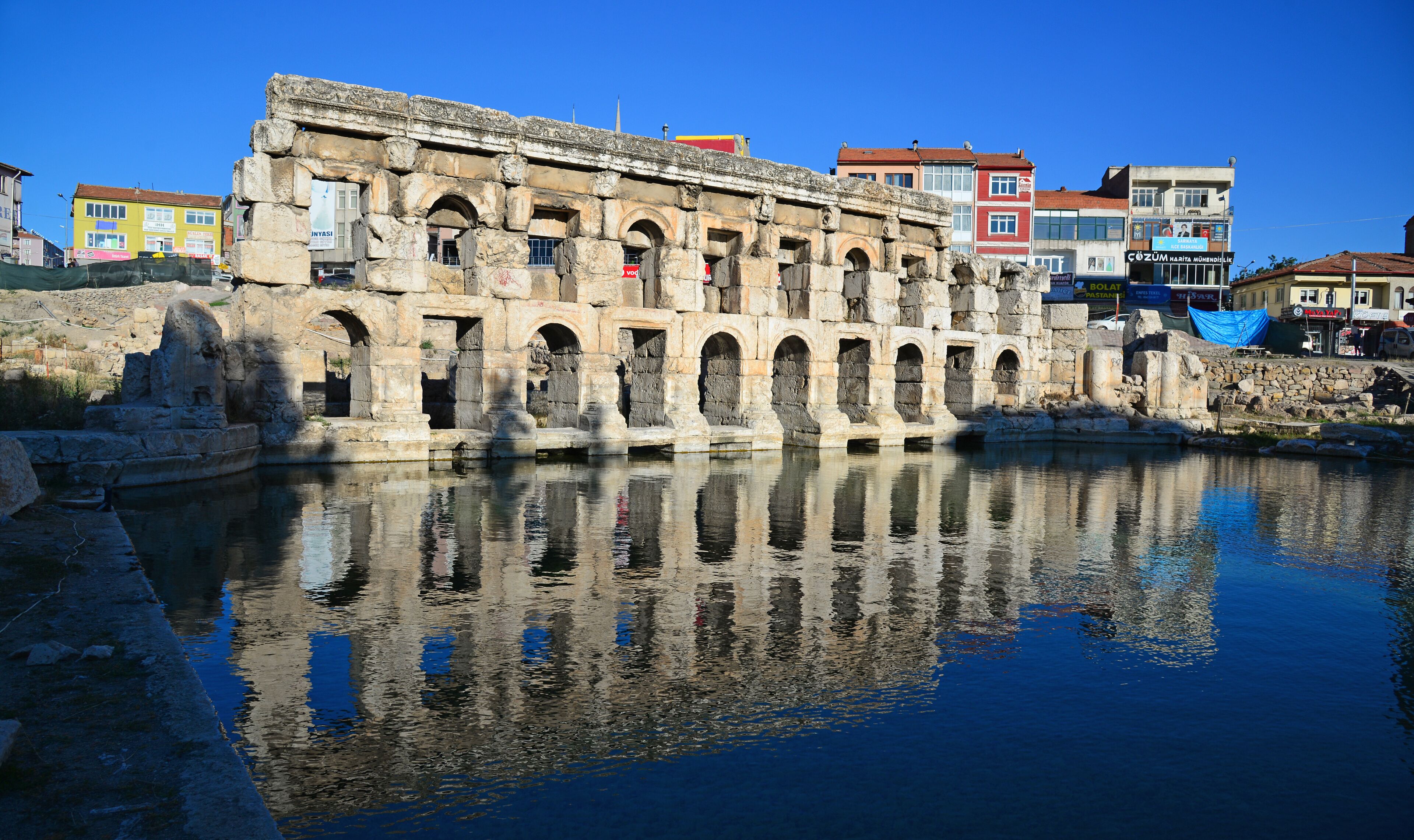 An ancient stone structure with arches reflecting in the water, surrounded by modern buildings.