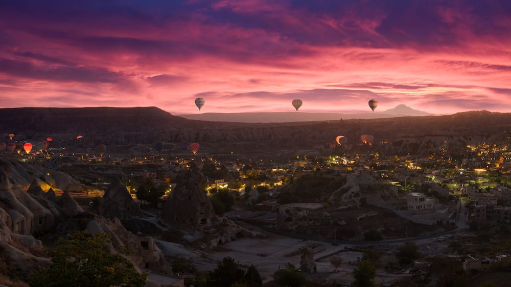 Ein Stadtbild im Abenddämmerung mit Heißluftballons und einem Bergpanorama im Hintergrund.