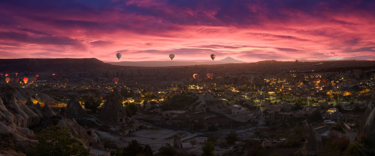 A cityscape at dusk with hot air balloons and a mountainous backdrop.