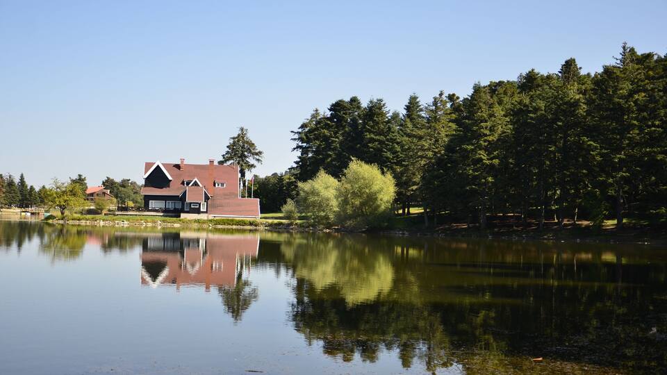 A house by a calm lake with reflections, surrounded by trees and a clear sky.