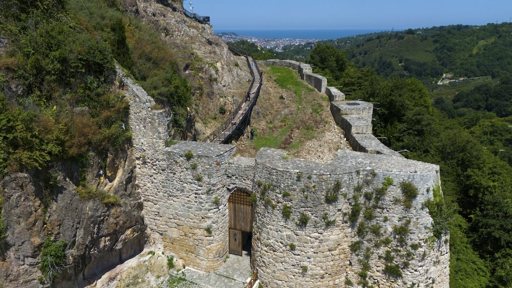 Une fortification en pierre dotée d’une porte en bois, entourée d’une végétation luxuriante et offrant une vue dégagée sur la côte au loin.