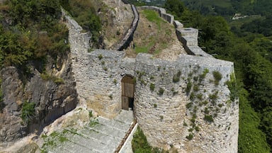 A stone fortification with a wooden door, surrounded by lush greenery and a clear view of the coastline in the distance.
