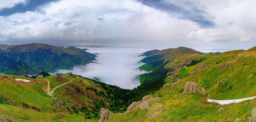 Un valle con una carretera, colinas verdes y una cadena montañosa cubierta de nubes.