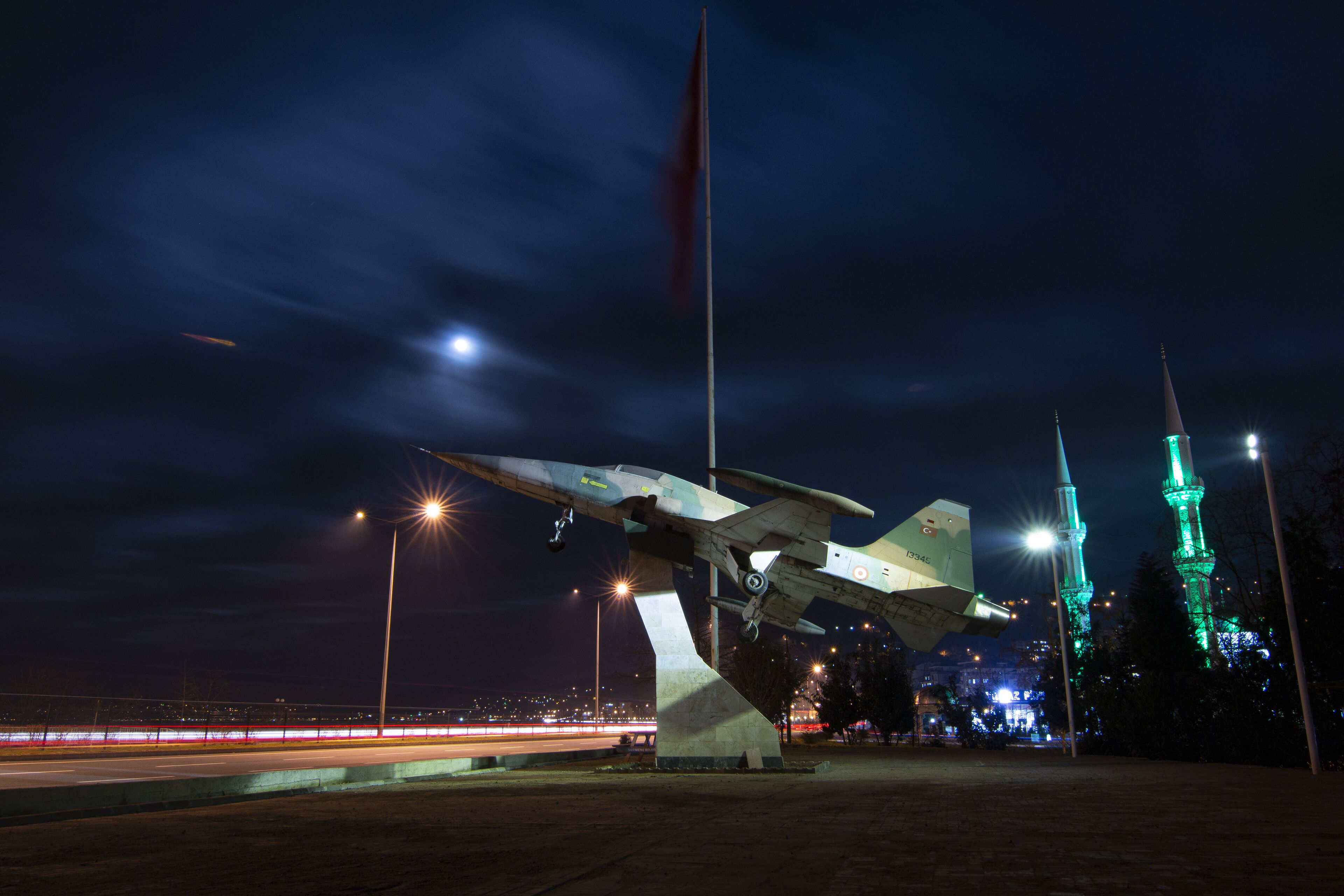 A nighttime sculpture of a military aircraft with a flagpole, illuminated by streetlights, and two green-lit towers in the background.