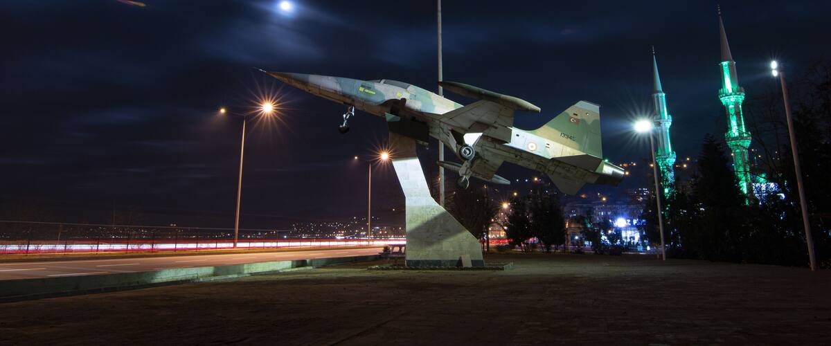 A nighttime sculpture of a military aircraft with a flagpole, illuminated by streetlights, and two green-lit towers in the background.
